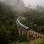 Blue train crossing the iconic Nine Arches Bridge in Ella, Sri Lanka, surrounded by lush green forest and morning mist.
